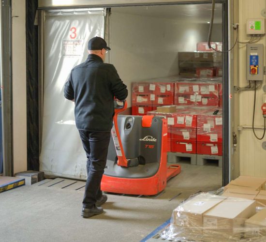 Ein Mann in schwarzer Kleidung und Mütze bedient einen roten Linde-T15-Elektrohubwagen in einem Lagerraum, um Paletten mit rot eingepackten Kartons zu bewegen.