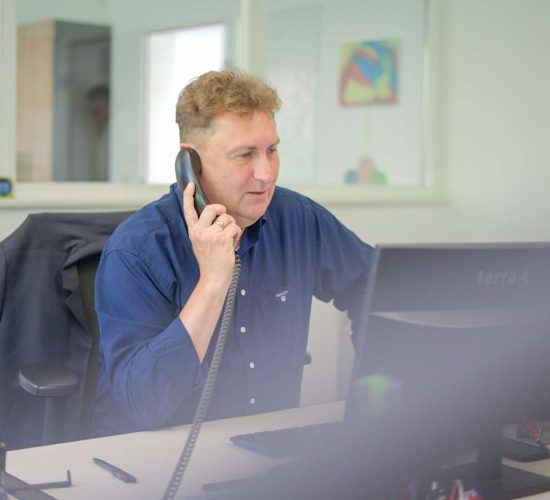 Ein Mann mit blauem Hemd sitzt an einem Schreibtisch und spricht am Telefon, während er auf einen Computerbildschirm schaut; im Hintergrund Bürofenster und ein Jackett auf dem Stuhl.