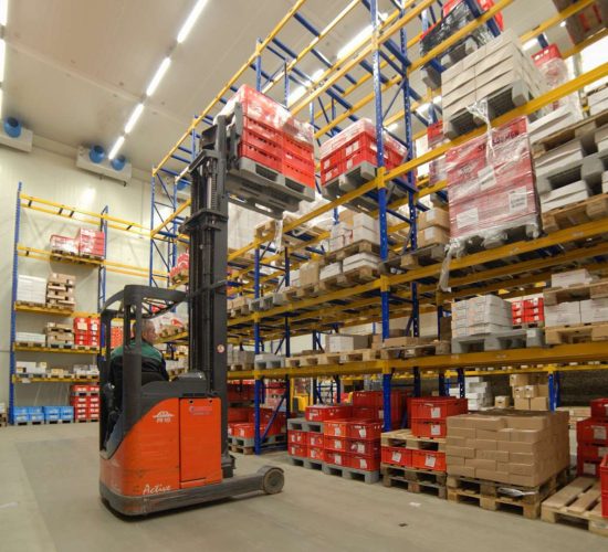 Gabelstapler hebt rote Kisten auf Paletten in einem großen Lager mit hohen Regalen voller Kartons und Kunststoffboxen