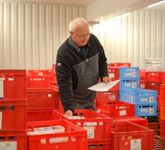 Ein älterer Mann mit Brille und schwarzen Pullover sortiert in einem Lagerraum rote und blaue Kunststoffkisten und hält dabei ein weißes Blatt Papier in der Hand.