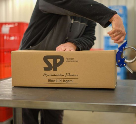 Person verpackt einen Karton mit Aufdruck "SP Spezialitäten Partner Bitte kühl lagern!" auf einem Metalltisch in einem Lagerraum mit roten und blauen Kunststoffbehältern im Hintergrund.